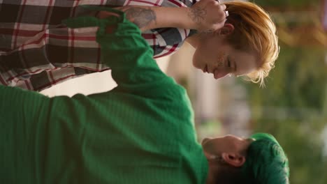 Vertical-video:-a-girl-with-green-short-hair-in-a-green-shirt-straightens-the-hair-of-her-blonde-short-haired-girlfriend-with-sparkles-on-her-face-in-a-plaid-shirt-and-chatting-with-her-during-her-date