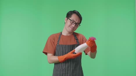 ama de llaves masculina asiática con un delantal mirando el spray en la mano luego mostrando los pulgares hacia abajo gesto a la cámara en el estudio de pantalla verde