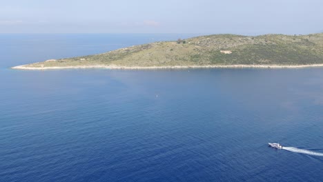 Drone-view-in-Albania-flying-over-blue-clear-ocean-with-a-small-green-island-and-a-small-boat-passing-by-on-a-sunny-day