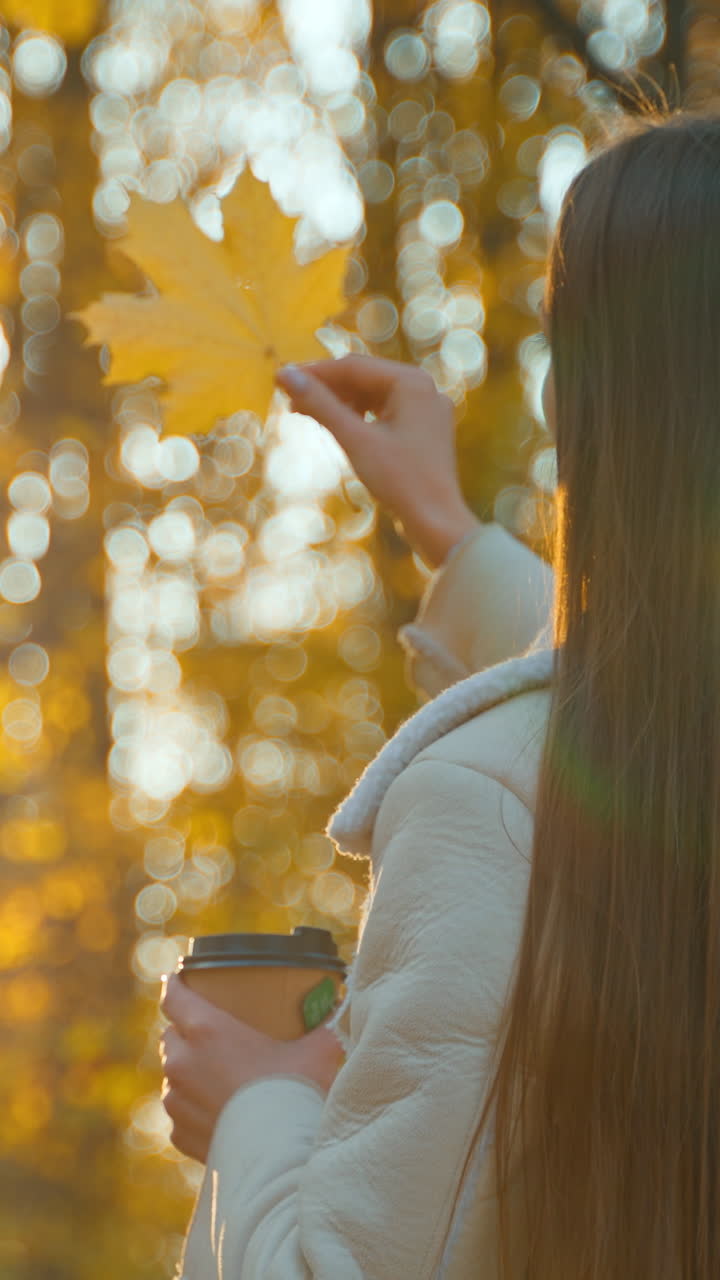 Long-haired brunette standing her back to camera against sun. Lady holding a paper cup in one hand and maple leaf in another. Autumn park backdrop in blur.