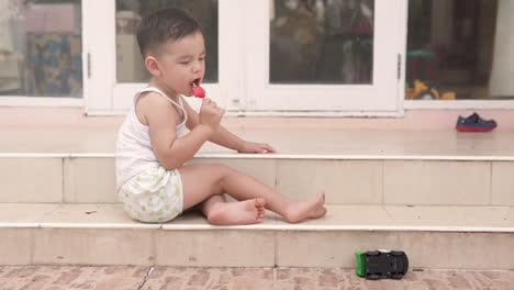 slow motion video of a handsome asian kid from the side eating a posicle ice cream sitting on the stairs of a house in summer