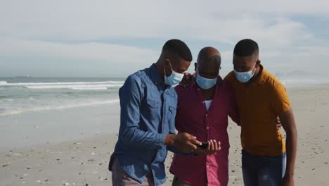 african american father and twin teenage sons wearing face masks taking selfies on beach