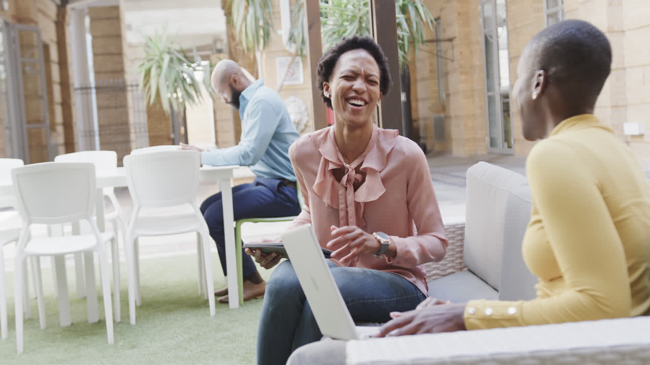 Two happy african american businesswomen talking, laughing and using laptop outdoors, in slow motion