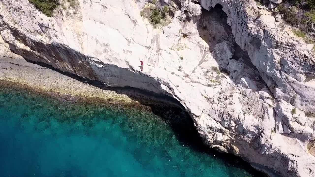 vista de pájaro de dron de un hombre parado en un acantilado y saltando haciendo una voltereta frontal, luego el dron se aleja