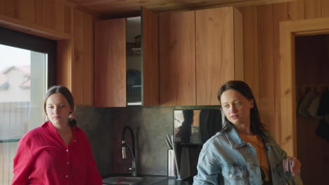 woman in denim outfit reaches up to retrieve glass plate from wooden kitchen cabinet while two female friends stand nearby, one watching by window and another rising from seat to assist