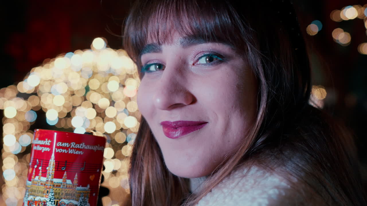 primer plano de una hermosa mujer de cabello oscuro diciendo saludos con su mano y bebiendo ponche de la taza de corazón rojo en el mercado navideño de viena, rodeada de luces navideñas en el fondo