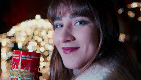 close up of a beautiful woman with dark hair saying cheers with her hand and drinking punch from red heart cup at vienna christmas market , surrounded by christmas lights in the background