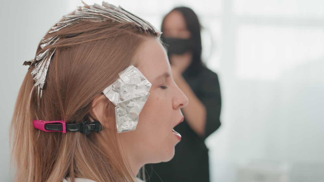 Donna con capelli trattati in un salone, colorazione e applicazione di stagnola in corso, parrucchiere che fotografa sullo sfondo, cliente che reagisce a bocca aperta, primo piano del profilo laterale, stagnola argentata e clip di sezionamento, delicato