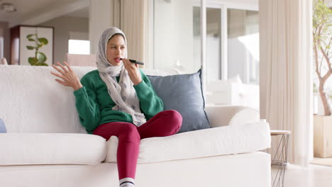 Biracial-woman-in-hijab-using-smartphone-on-sofa-at-home-with-copy-space,-slow-motion