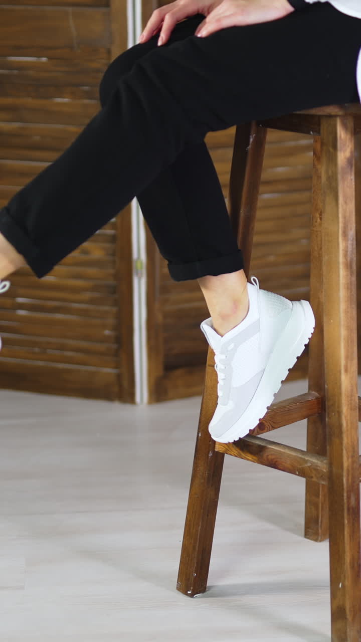 Woman sits on a high stool waving her feet in white sneakers. Model jumps off the tabouret and walks away in comfortable footwear. Vertical video
