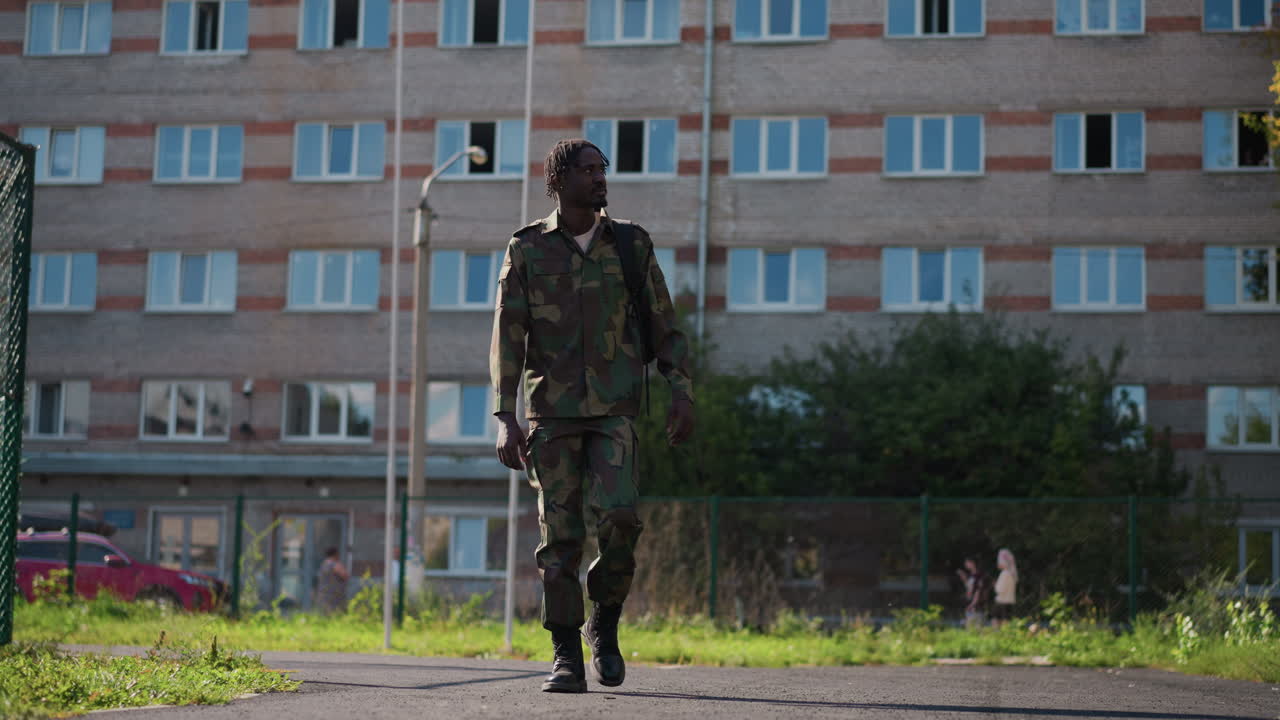 Uniformed Figure Advances Purposefully Through City Landscape With Determined Stride, Courageous Soldier Moves Steadily Towards Camera Amid Modern Apartment Buildings And Playground Scenery