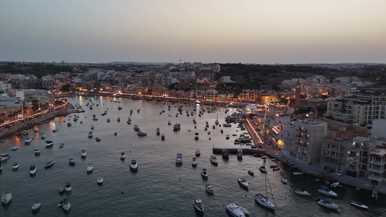 Flygvy över Marsaskala-bukten, Malta, med hamn, förtöjda båtar, lägenheter vid vattnet och festliga kvällsljus som reflekteras på det lugna vattnet under ett byfirande.