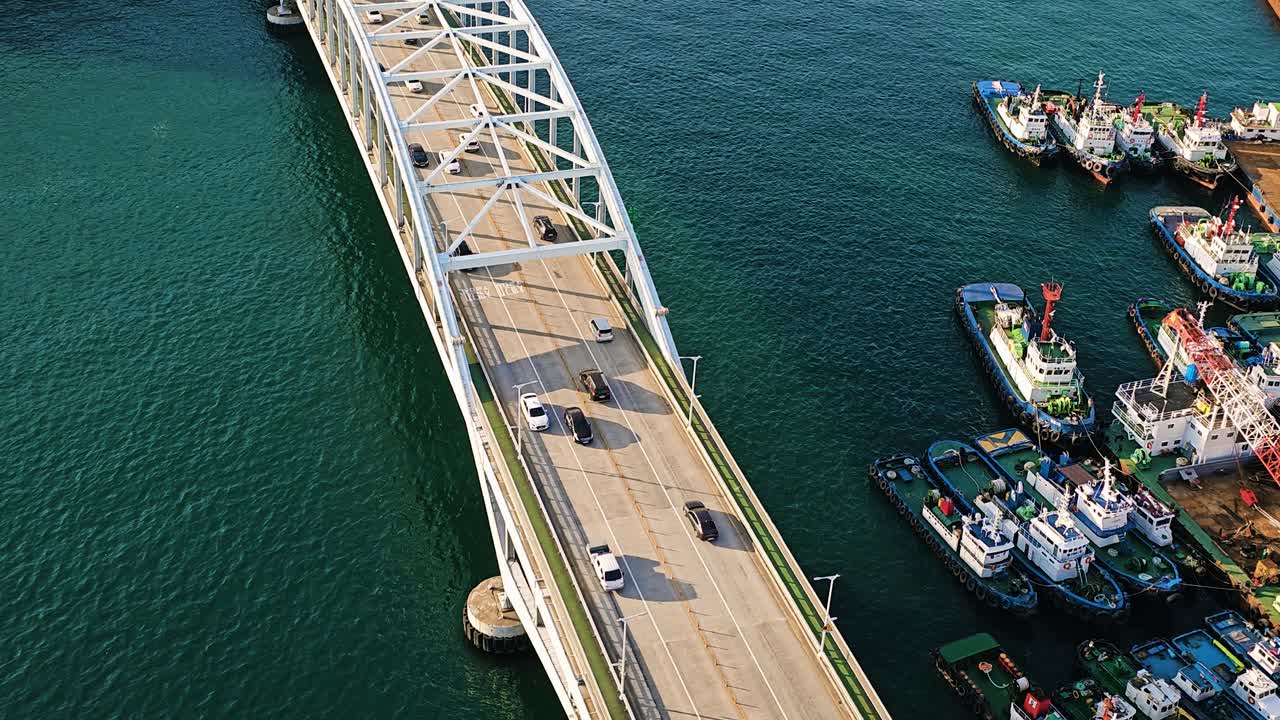 Aerial shot of Busan Bridge in South Korea showing moving traffic over blue ocean waters with nearby docked ships
