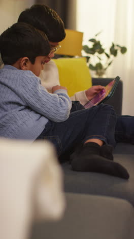 vertical video of two young boys sitting on sofa at home playing games or streaming onto digital tablet together 3