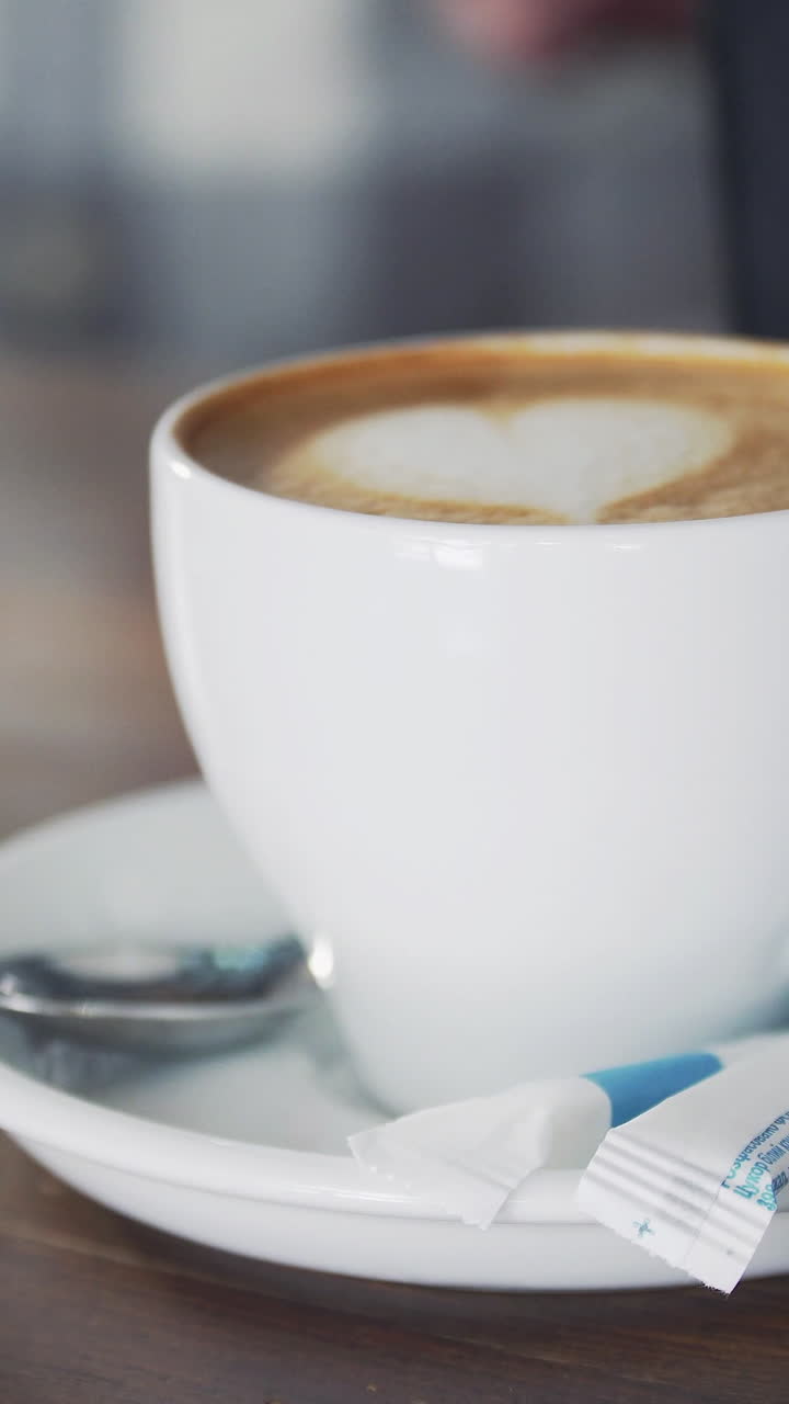 White cup with ready cappuccino standing on wooden table and barman is pouring milk into another cup. Vertical video.