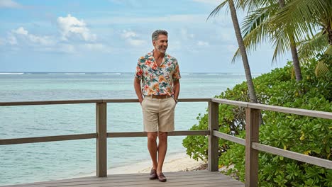 a relaxed man in a tropical shirt stands on a deck overlooking a beautiful ocean view surrounded by palm trees and lush greenery under a bright sky