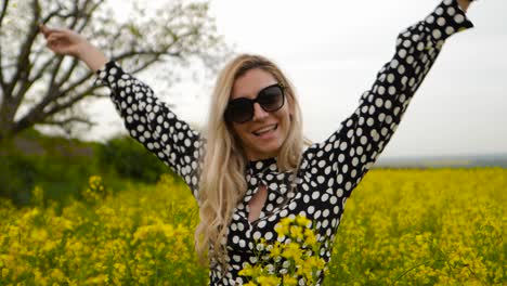 happy woman with sunglasses raises hands in canola field, flower in hand