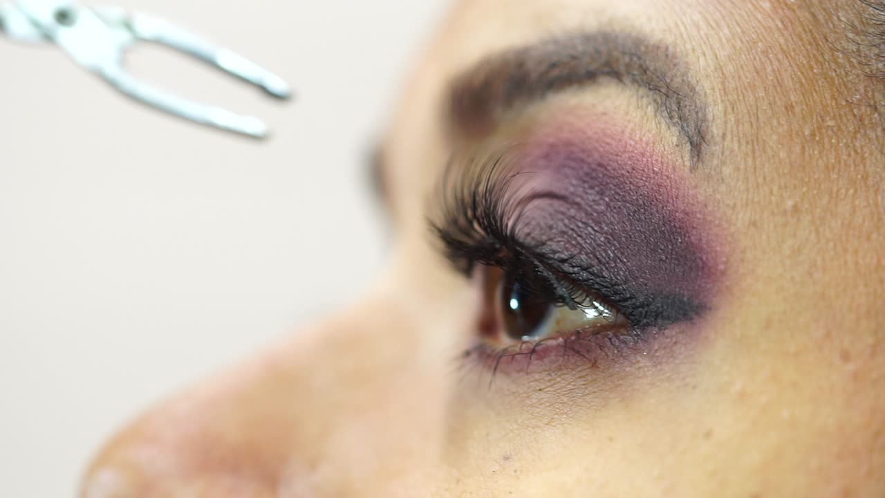 Side close-up of brown-eyed woman applying false eyelashes with tongs