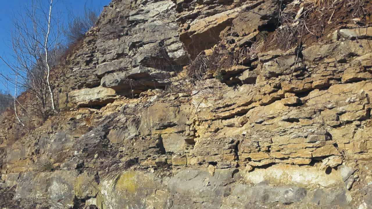 Sun shining on a crumbled and steep yellow, brown limestone cliff or mountains wall in Tabasalu during a beautiful clear day with blue sky. Plants are growing on the wall with roots hanging on cliff.