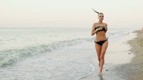 young attractive woman in black leather swimsuit running by the sea on the beach. her ponytail is waving in the wind while woman