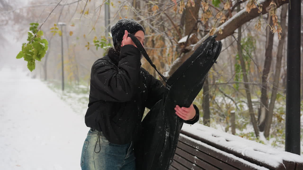 여자가 기타 을 제거하고, 눈으로 인 벤치에 놓고, 얼음으로 둘러싸인 조용한 겨울 공원 환경에서 눈이 부드럽게 떨어지는 동안 열고 있는 측면 시각