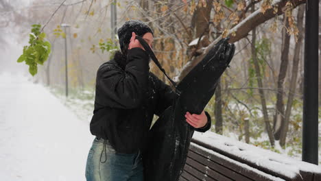 vista lateral de una mujer quitando el paquete de guitarra, colocándolo en un banco cubierto de nieve, y abriéndolo mientras la nieve cae suavemente en un tranquilo parque de invierno, rodeado de árboles helados