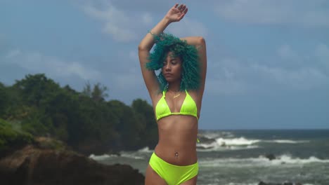 Young-girl-in-bikini-with-green-curly-hair-flowing-in-the-wind-on-a-cliff-side-beach