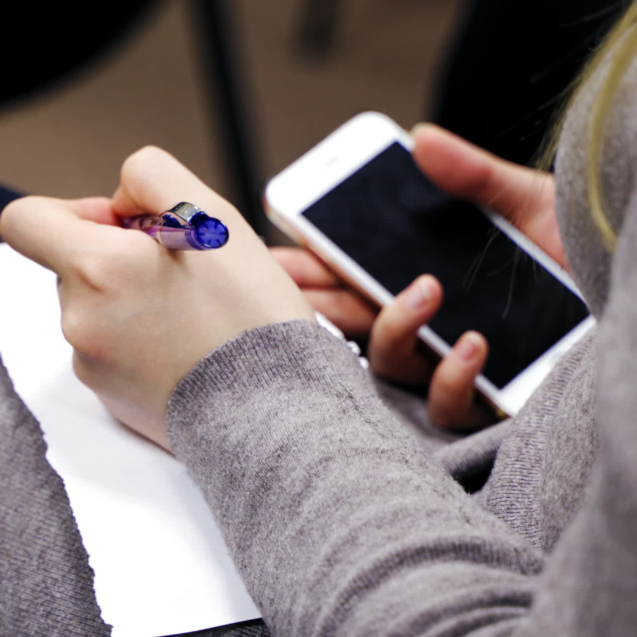 Woman is sitting and writing with a pen on a sheet of paper. Business woman holding phone and writes important information on paper during the conference indoors.