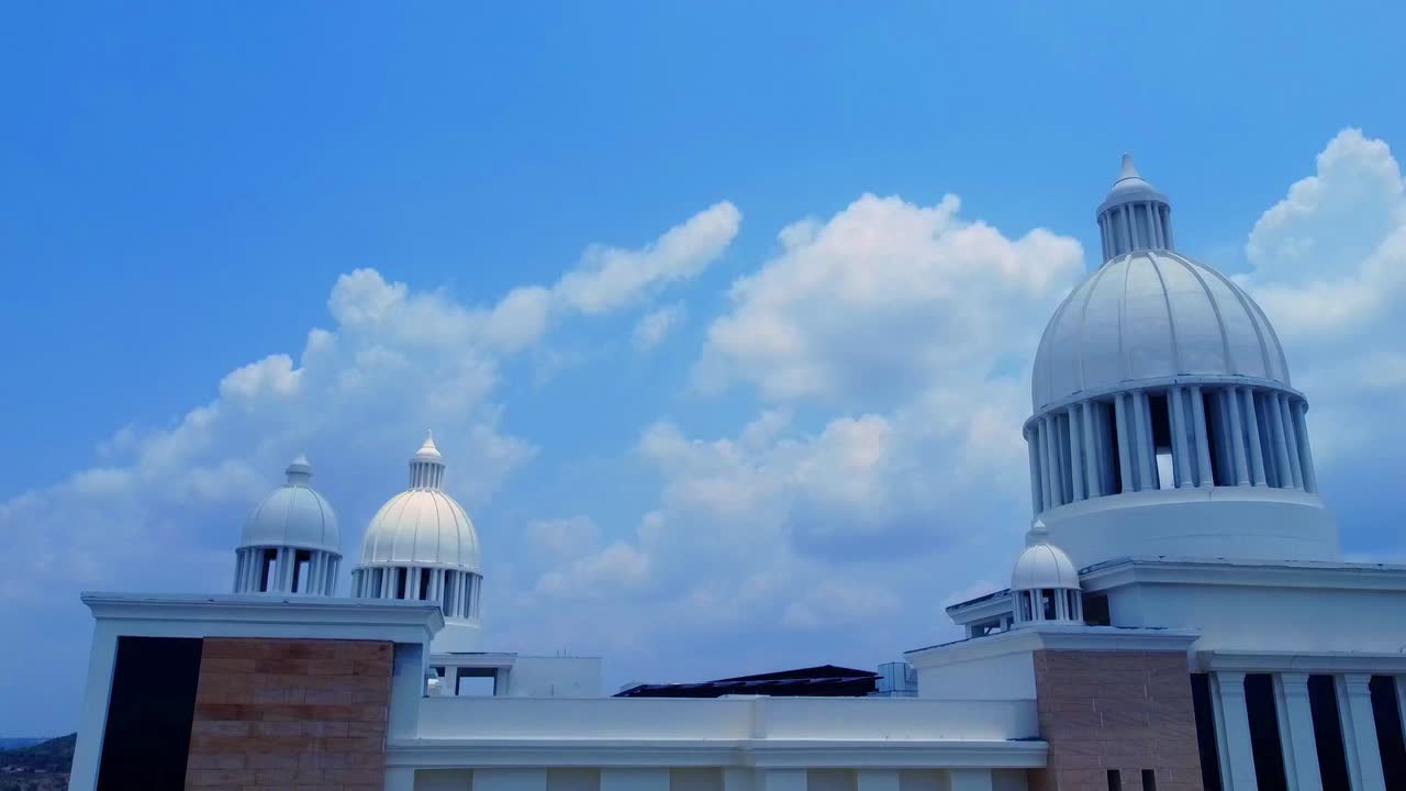 Ascending aerial shot revealing the grand, neo-classical architecture of the Kanha Shanti Vanam main building in India, its white domes majestic against a bright blue sky with fluffy white clouds