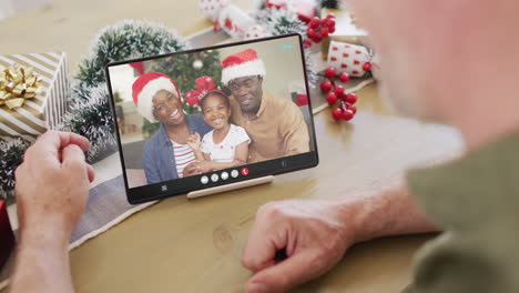 hombre caucásico con decoraciones de navidad teniendo llamada de video de tableta con una feliz familia afroamericana