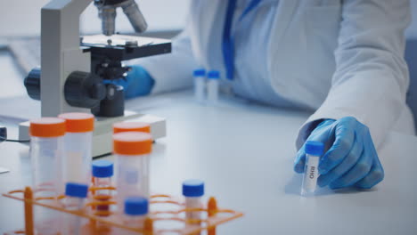 Close-Up-Of-Lab-Worker-Conducting-Research-Using-Microscope-Holding-Test-Tube-Labelled-Rho