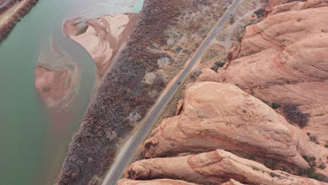 Vista-Aérea-Cinematográfica-Del-Vehículo-Oscuro-Que-Se-Mueve-En-La-Carretera-En-El-Paisaje-Desértico-De-Utah-Por-El-Río-Colorado-Bajo-Altas-Colinas-Empinadas-De-Arenisca,-Disparo-De-Drones-De-Alto-ángulo-De-Arriba-Hacia-Abajo