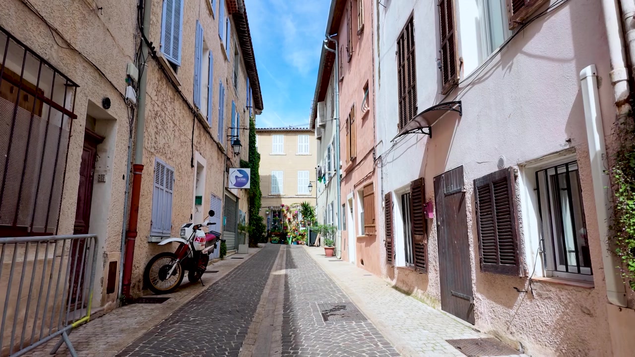 Una telecamera a bassa angolazione scivola attraverso un tranquillo vicolo mediterraneo illuminato dal sole, con edifici color pastello e persiane.