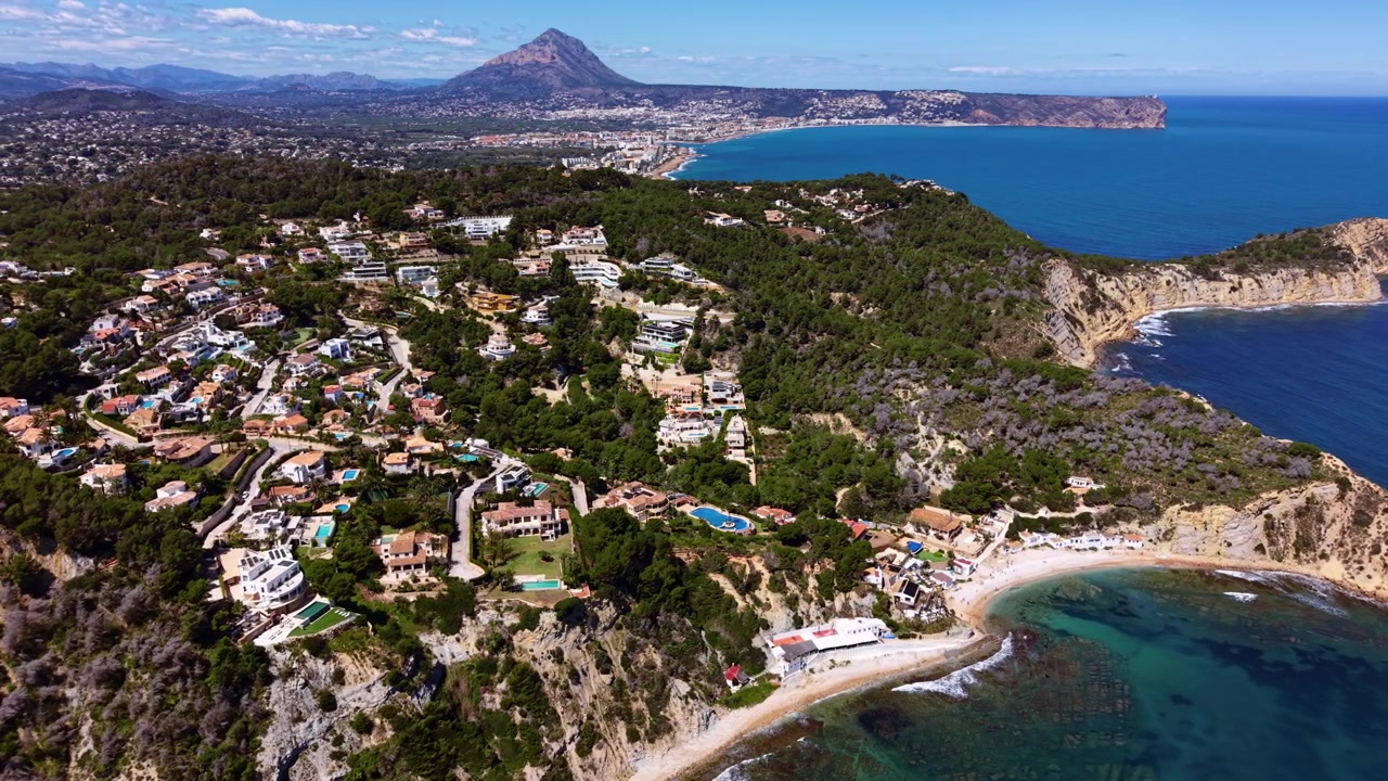 Litoral da Costa Blanca mostrando vilas de luxo aninhadas entre pinheiros em falésias rochosas acima das águas turquesa do Mediterrâneo, com a montanha Montgó ao fundo.
