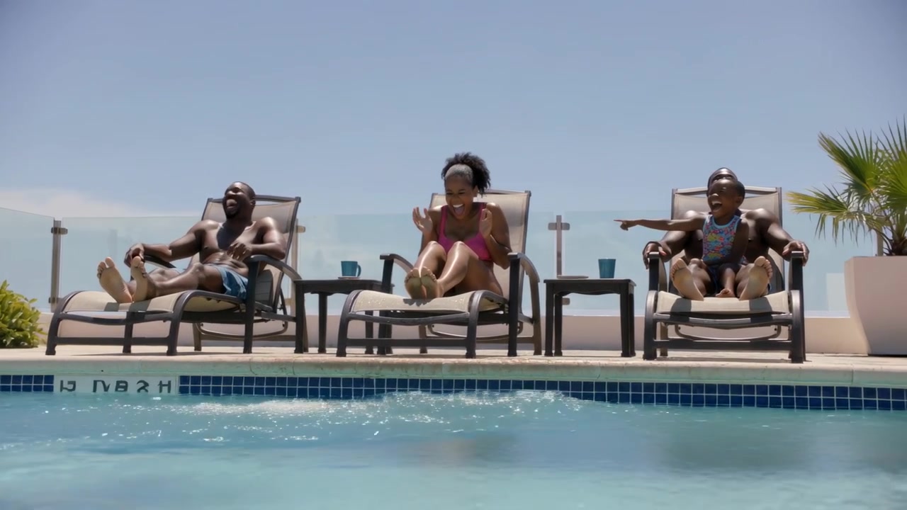 Kicking family on pool loungers splashing pool water, woman pink one-piece laughing, child pointing