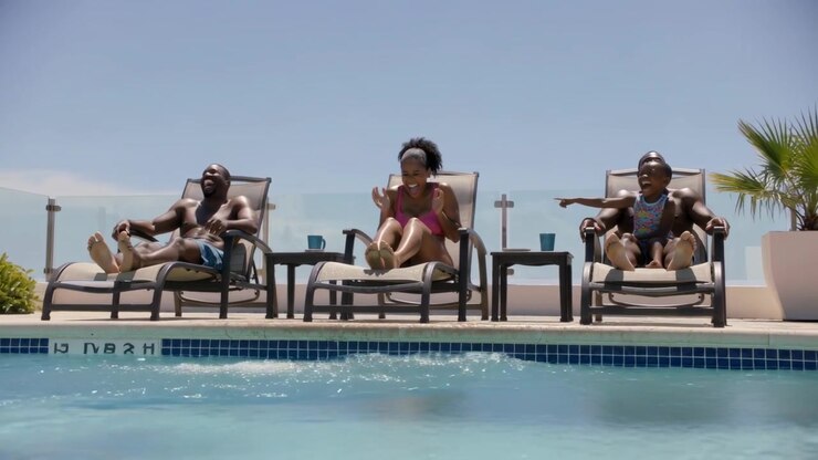 Kicking family on pool loungers splashing pool water, woman pink one-piece laughing, child pointing