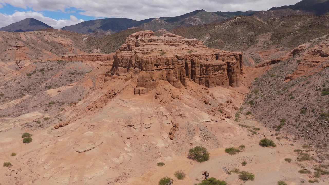 Aerial drone rises and pulls back from a towering eroded red sandstone formation near Angastaco in the Calchaqui Valleys, Salta, Argentina, revealing a dry riverbed and distant mountains.
