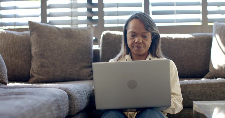 Mature African American woman positioning laptop and typing while working on sectional sofa at home