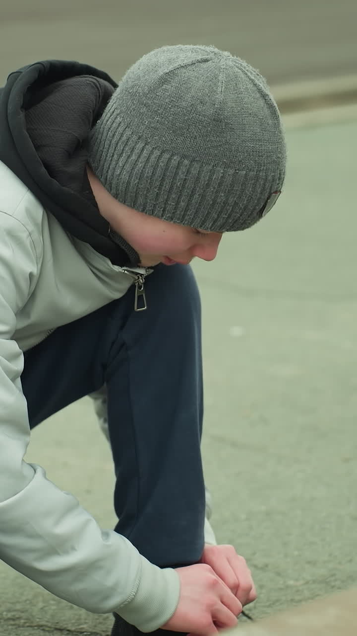 A young boy kneeling down tying his shoelace on a paved road, wearing a gray beanie and jacket, the background is slightly blurred with a minor pothole
