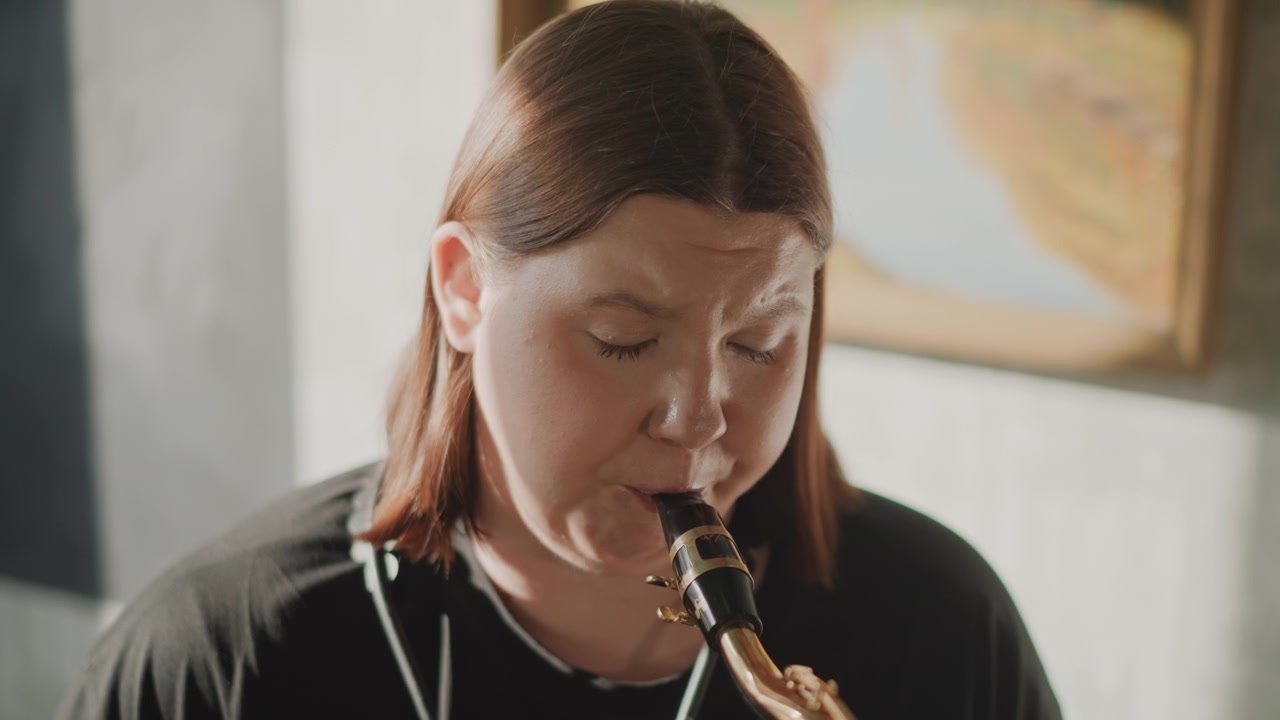 Sassofonista donna bianca che si esercita in un'aula, luce naturale soffusa, primo piano del viso, controllo del respiro e imboccatura, focus sull'imboccatura, atmosfera di coaching dell'insegnante, ambiente di scuola di musica, gradazione di colore calda.