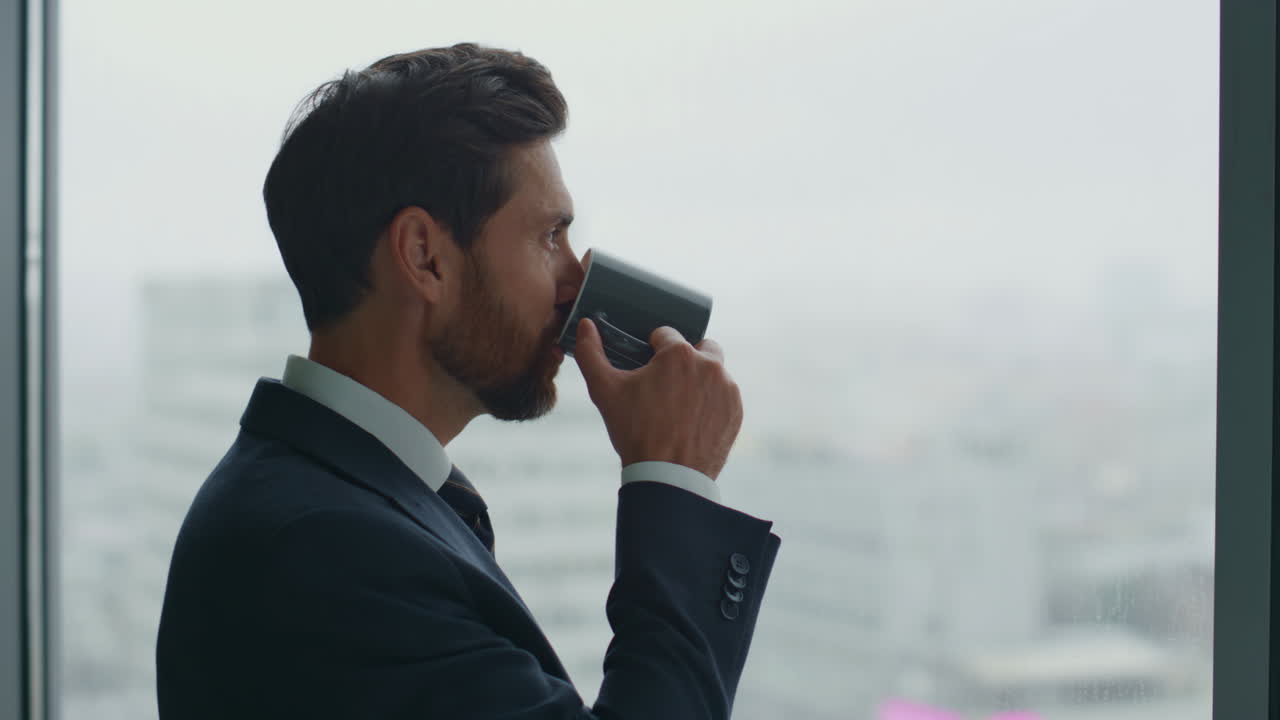 ceo sonriente bebiendo té mirando por la ventana de la oficina sintiendo el éxito de cerca.