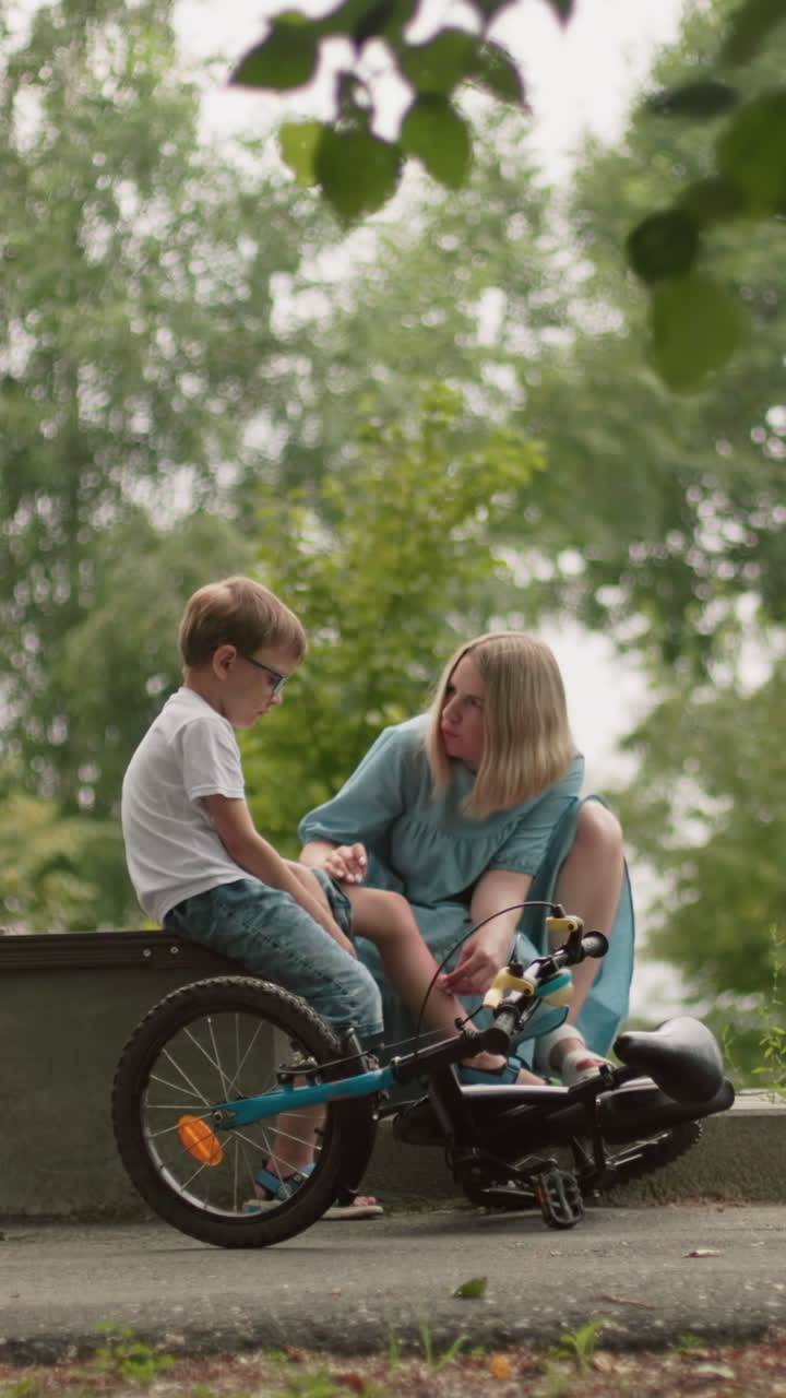 una madre atiende suavemente la herida de su hijo mientras está sentada a su lado en un banco en un parque, rodeada de vegetación, el niño sostiene sus pantalones mientras su madre limpia cuidadosamente su lesión en la pierna