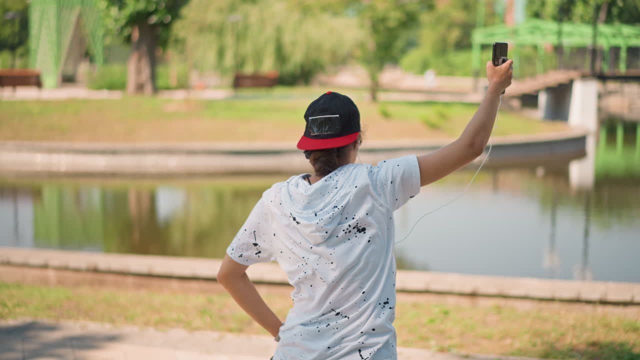 Bailarín de estilo libre en un parque junto a un estanque con teléfono y auriculares, joven con gorra practicando coreografía y gestos divertidos en un camino pavimentado junto al agua, ambiente de artista callejero, ensayo en una tarde soleada