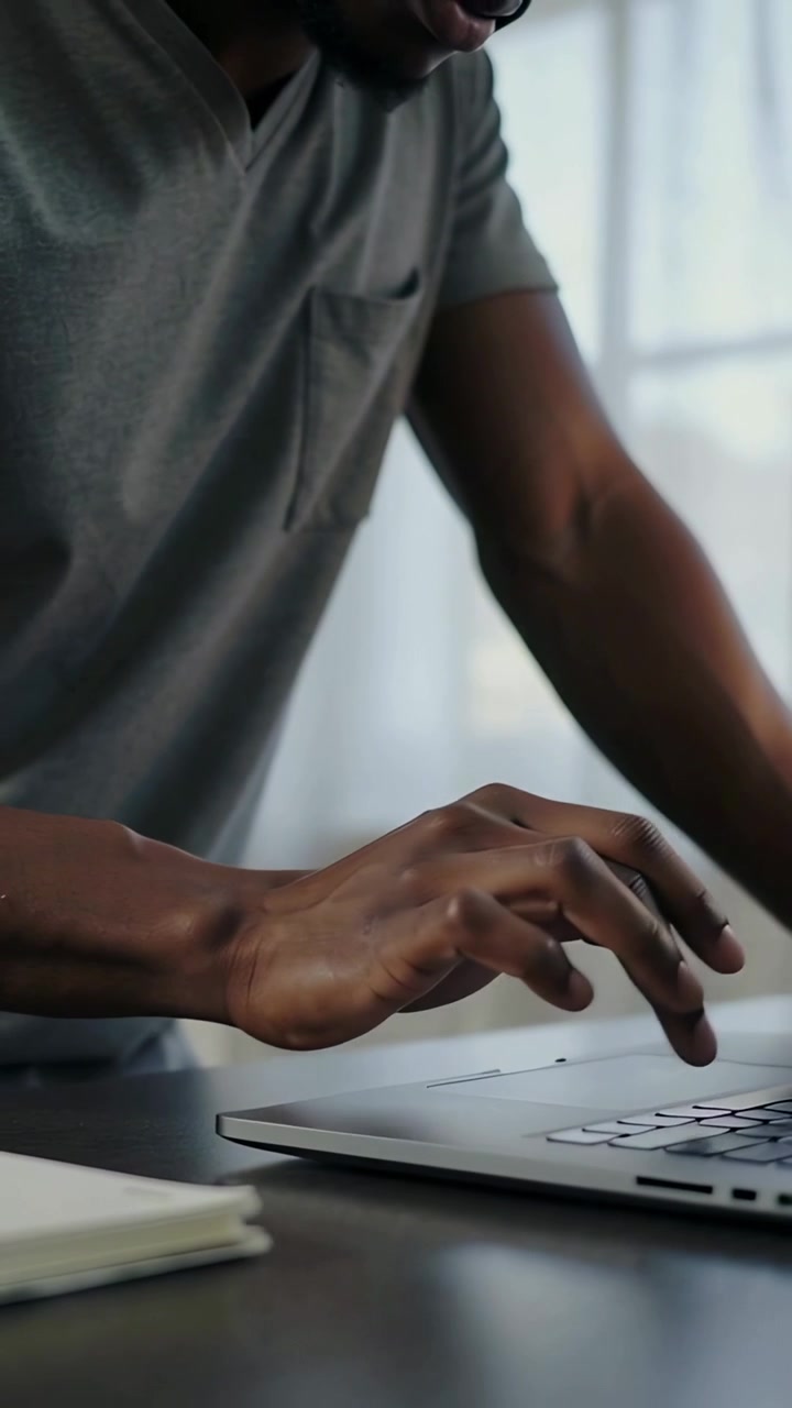 Vertical video: Leaning man in gray polo typing on laptop at desk, working with notebook and window