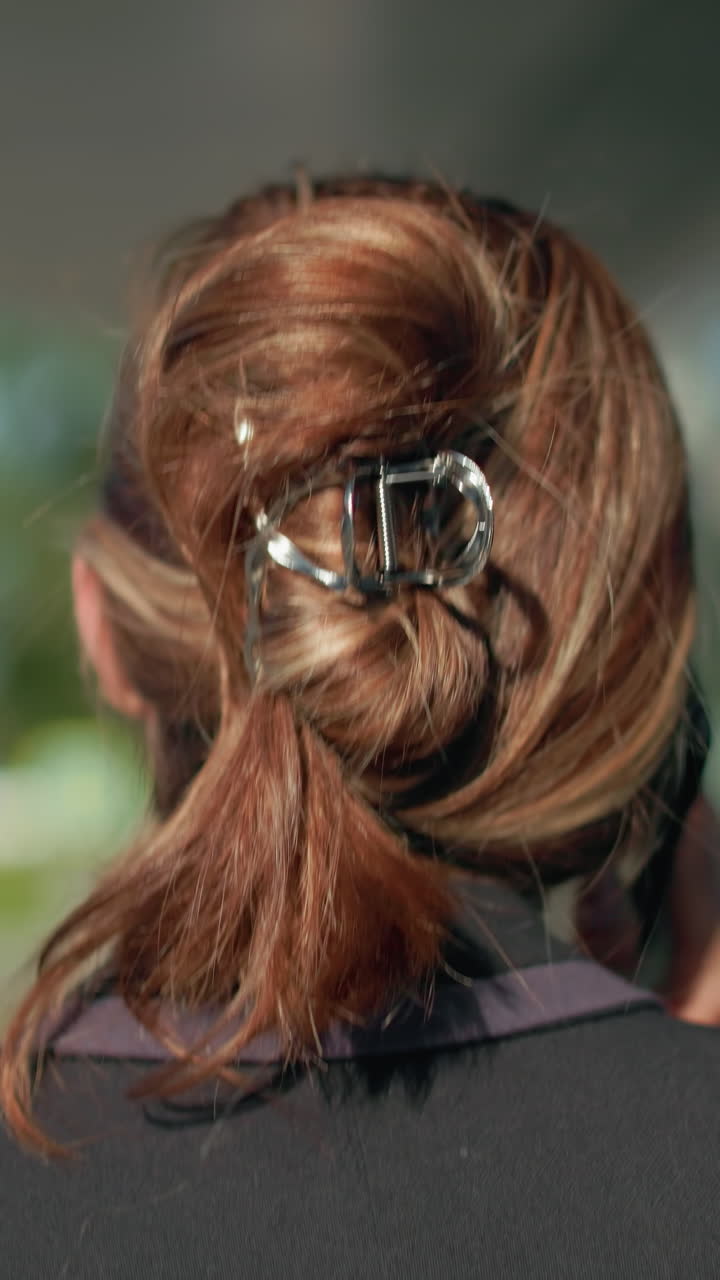 Close up rear view of professional woman in black suit with tied-back hair sipping coffee while on phone call walking near glass building with reflection of moving car in urban environment and soft sunlight