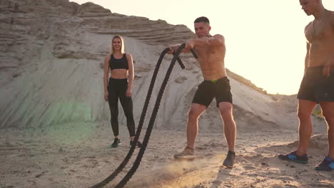 Crossfit-Grupal-Entrenando-A-Un-Hombre-Con-El-Torso-Abierto-Haciendo-Ejercicios-Con-Una-Cuerda,-Una-Mujer-Y-Un-Hombre-Observando-La-Actuación-En-Un-Sitio-Arenoso.