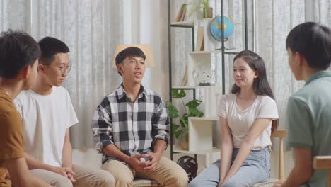아시안 십대 그룹은 의자에 앉아서 집에서 논의하는 원을 형성하고 있습니다. 모두가 카레드 셔츠를 입은 소년이 자신을 소개하는 것을 듣고 있습니다.
