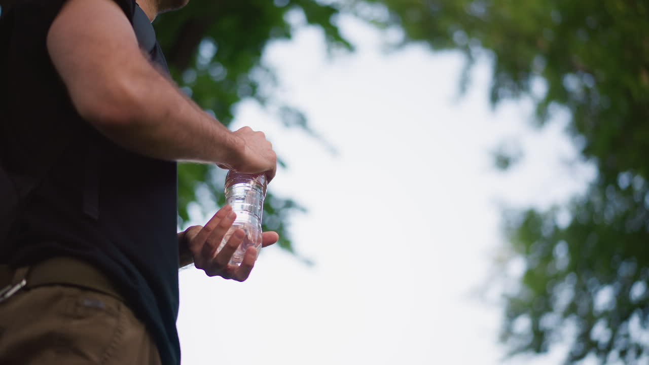 CloseUp Backpacked Caucasian Man Holding Bottle, Outdoor Park Scene With Clear Plastic Bottle, Leafy Canopy, Casual Black Shirt And Steady Grip, Quick Hydration Pause During Trail Walk