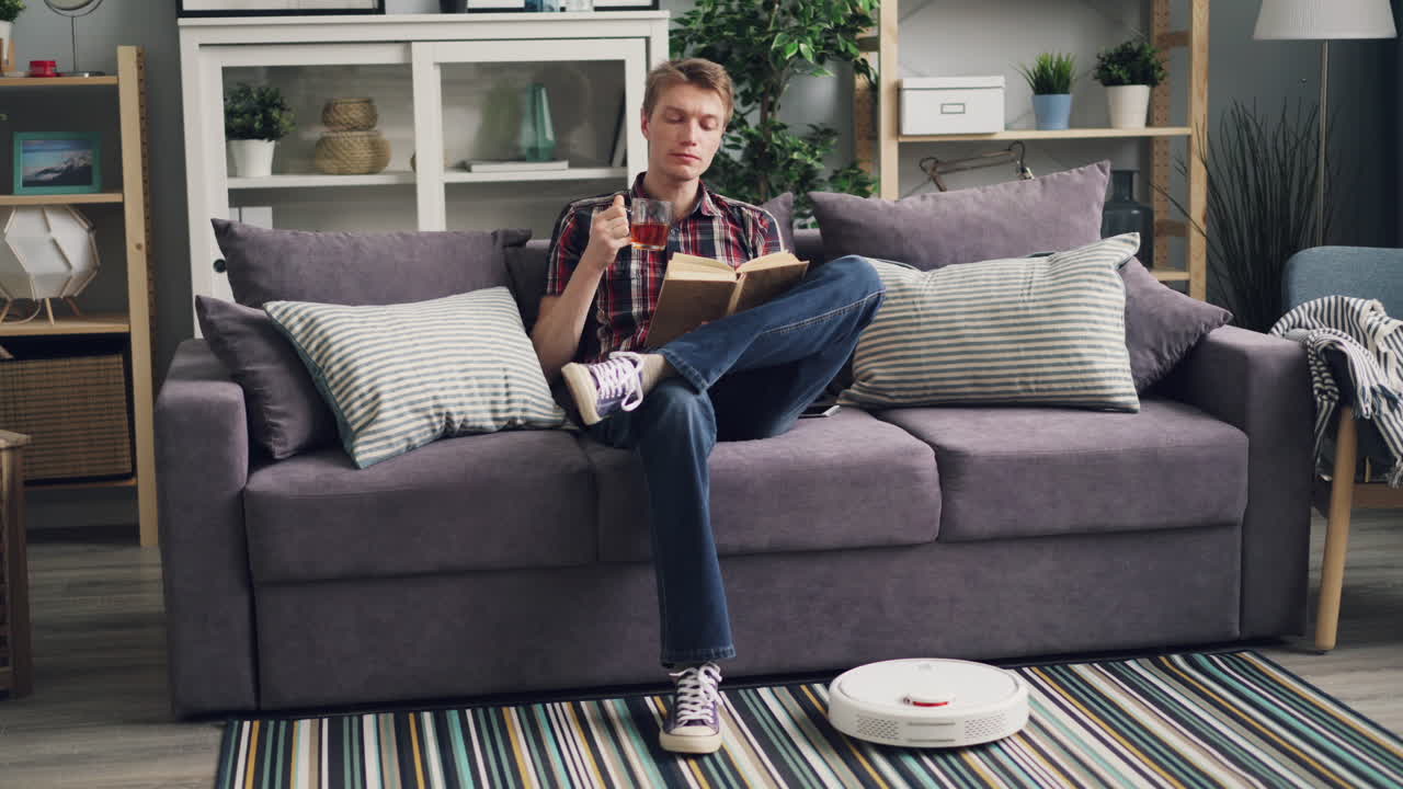 Man Relaxing on Sofa, Reading a Book, with a Cup of Tea, Robot Vacuum