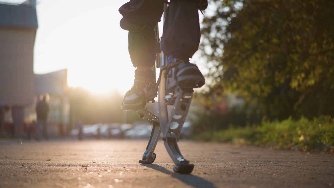어두운 바지와 스니커를 입고 교외 야외 지역에서 스프링 스을 사용하여 남자의 다리의 클로즈업. 배경은 흐릿한 나무, 건물 및 거리를 보여줍니다. 낮은 카메라 각은 움직임을 강조합니다.